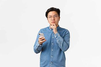 thoughtful creative asian man in glasses thinking while making post on social media, looking away, pondering or making decision, holding smartphone, choosing something in internet